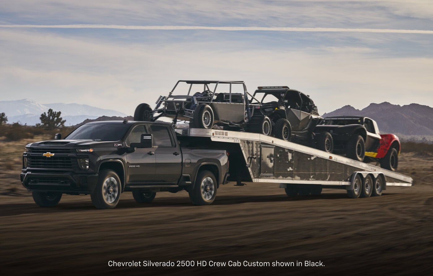 Front three-quarter view of a 2026 Chevrolet Silverado 2500 HD Custom Crew Cab in Black hauling a multi-car gooseneck trailer.
