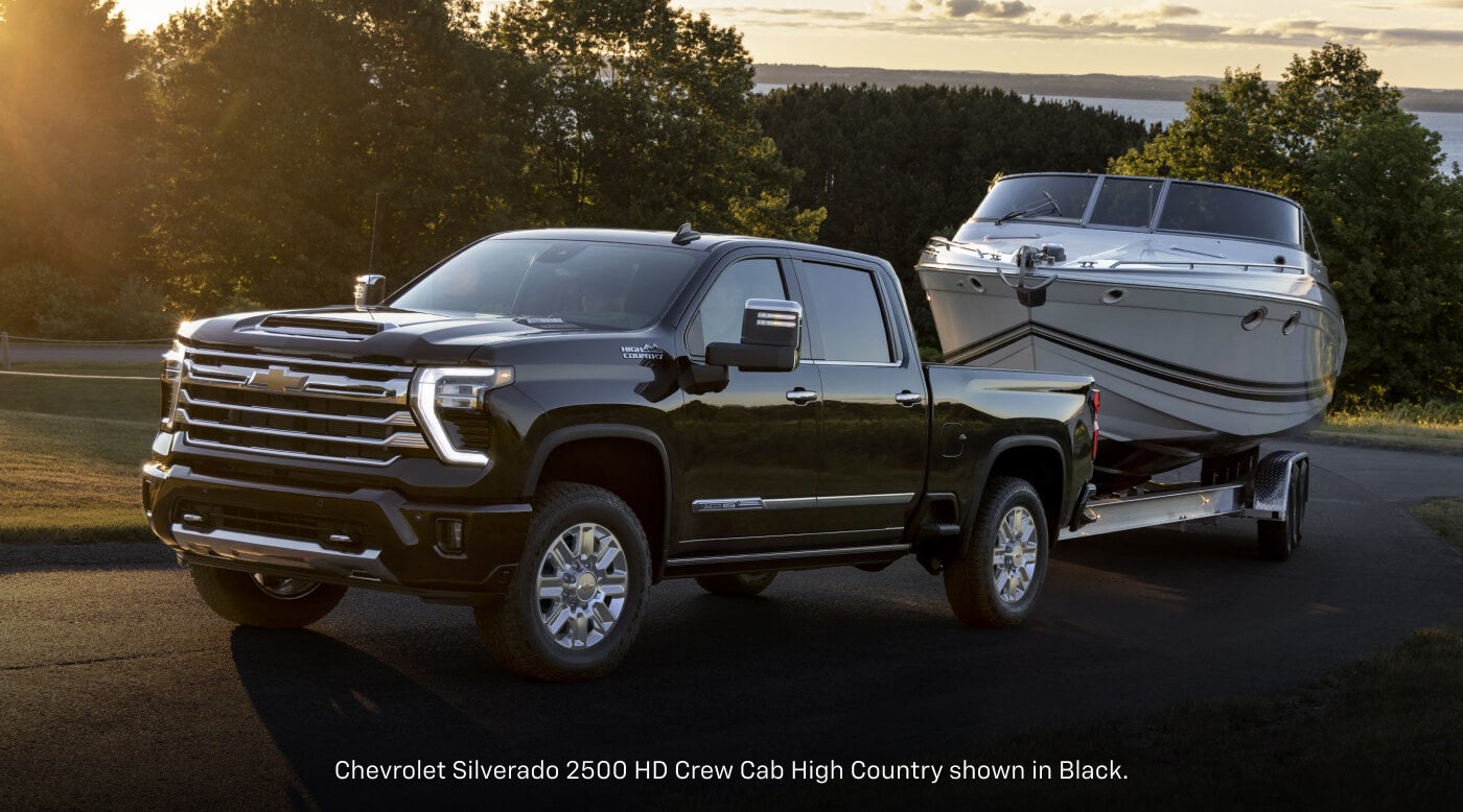 Front three-quarter view of a 2026 Chevrolet Silverado 2500 HD High Country Crew Cab in Black towing a large cabin cruiser boat.