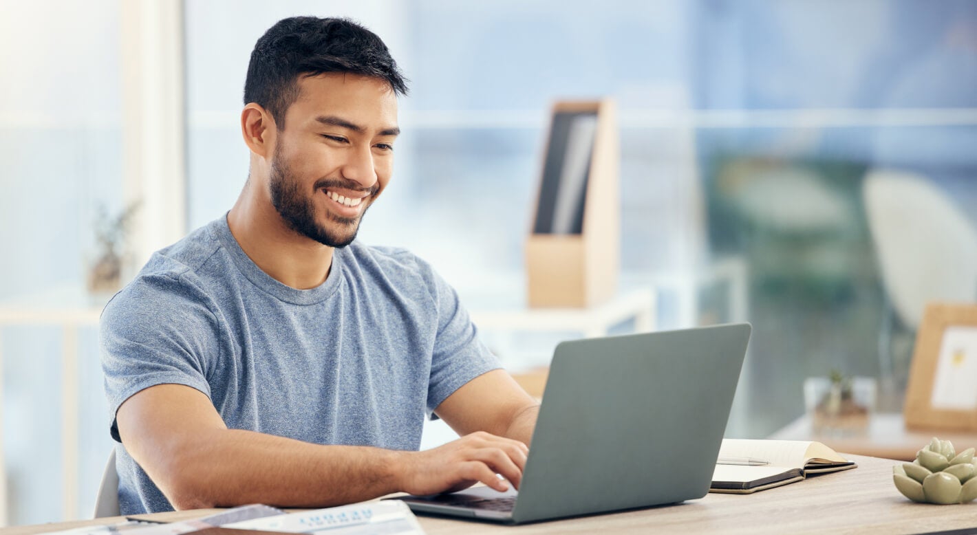 A man sits at a desk in a bright sunny room and smiles while using a laptop.