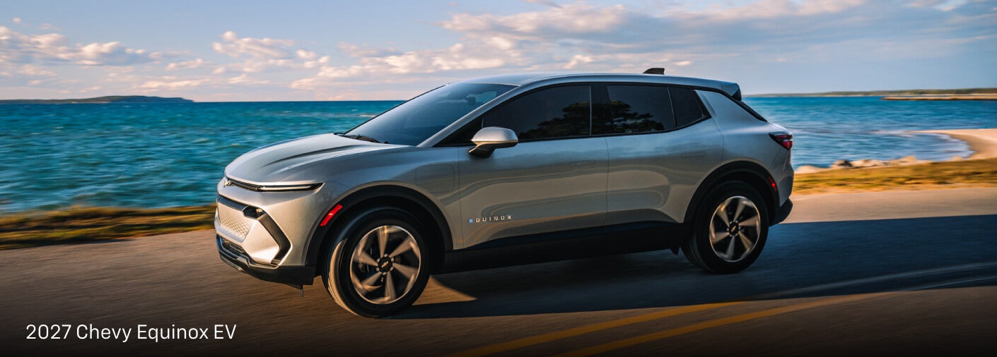 Exterior side action shot of a light blue Chevy Equinox EV driving down a seaside highway.